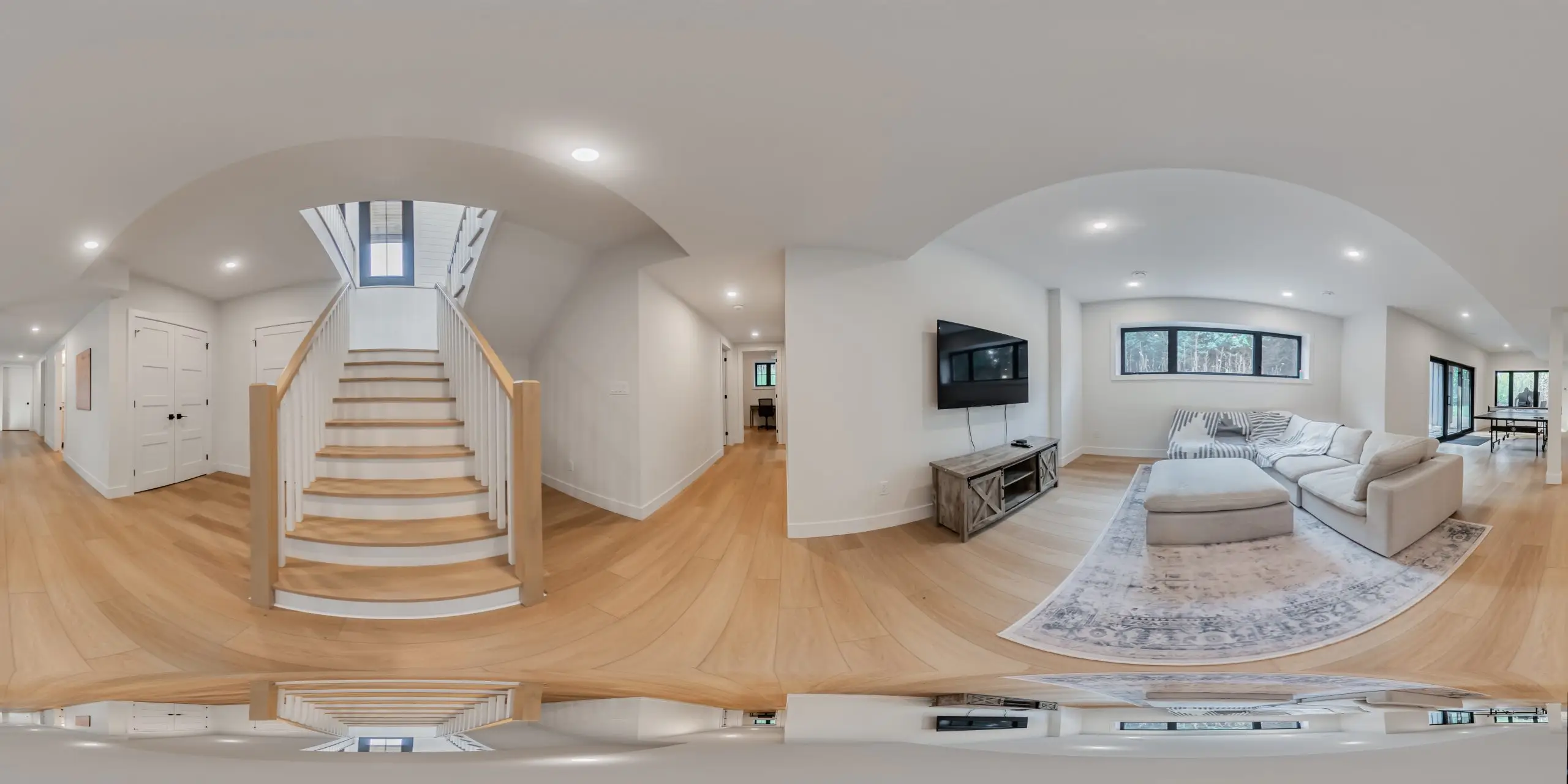Panoramic view of a bright modern home interior with a central staircase, hardwood floors, wall-mounted TV, sectional sofa, rug, and soft neutral décor.