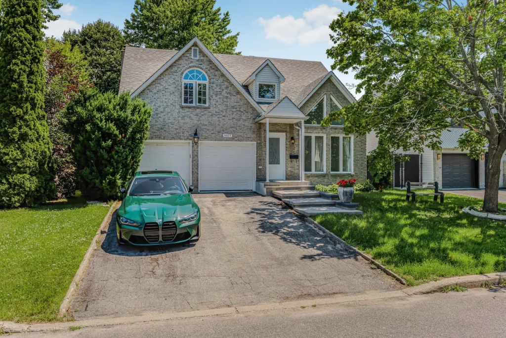 A green car is parked in the driveway of a two-story brick house with a two-car garage and a small front lawn with trees.