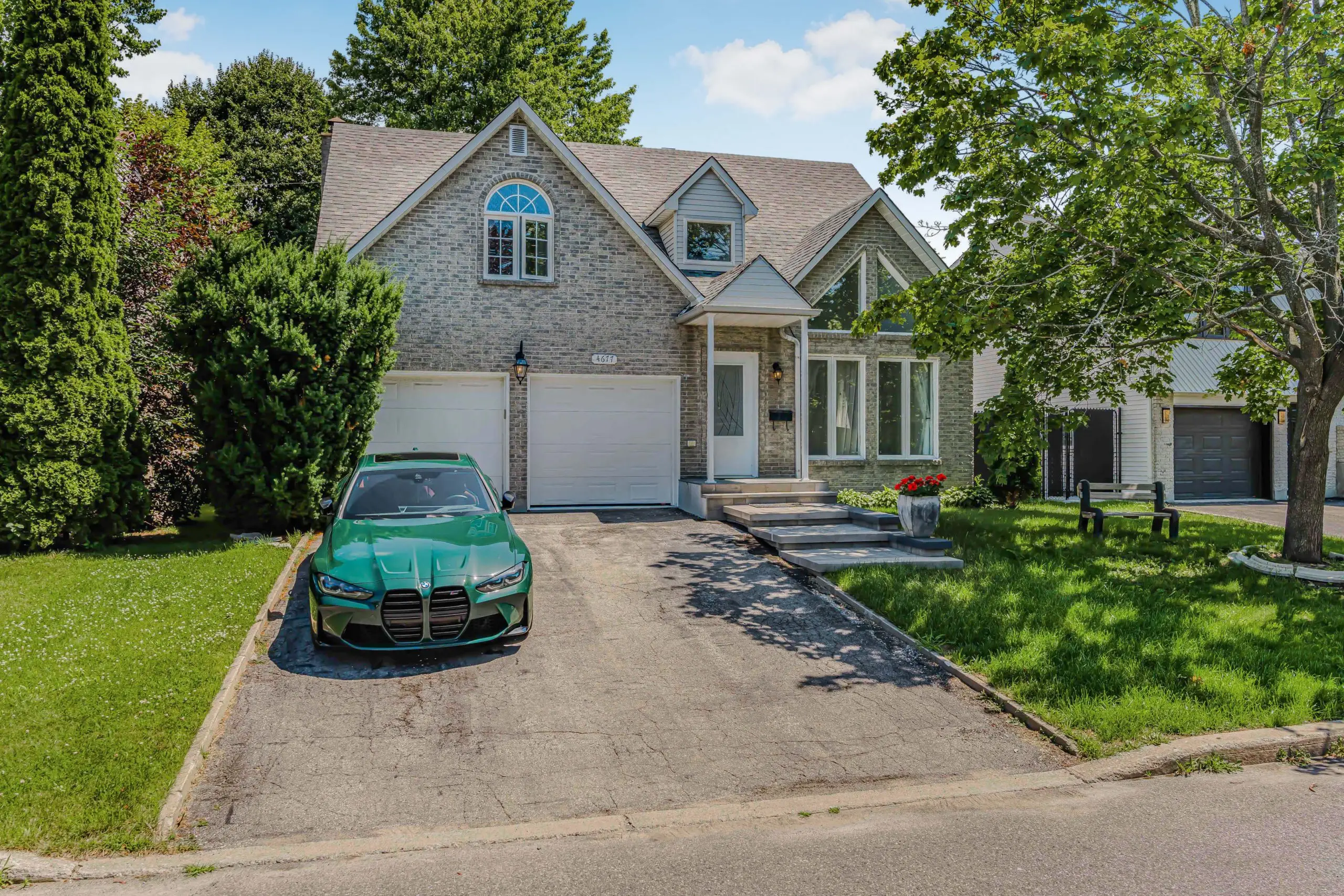 A green car is parked in the driveway of a two-story brick house with a two-car garage and a small front lawn with trees.